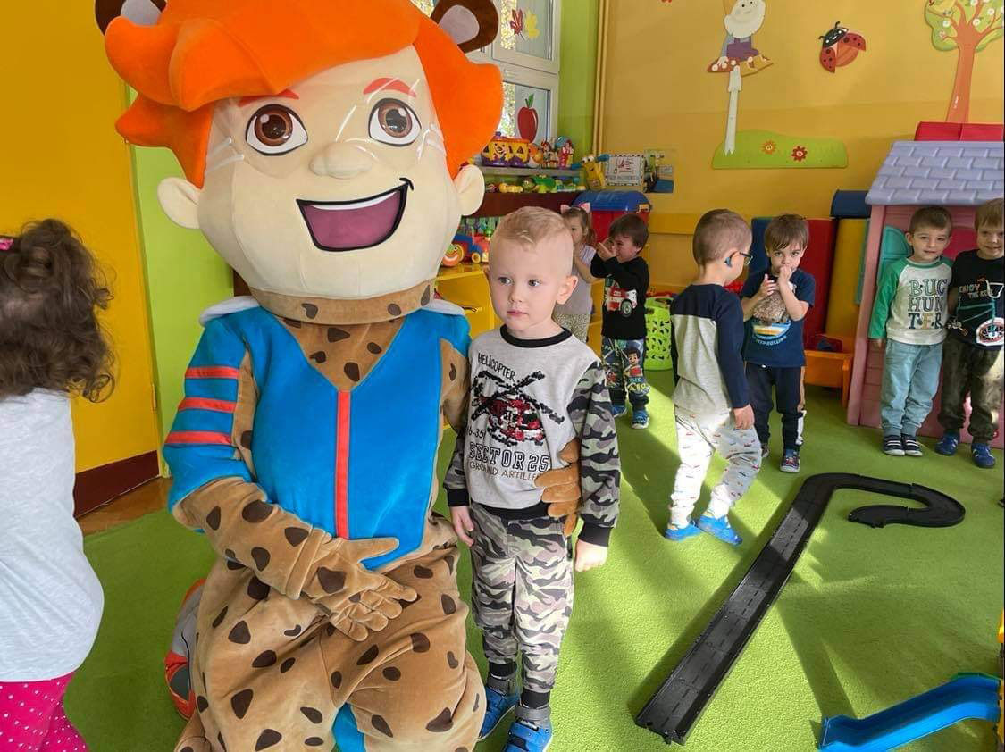 Na fotografii widnieje strój reklamowy w postaci chłopca pozujący do zdjęcia w towarzystwie dzieci